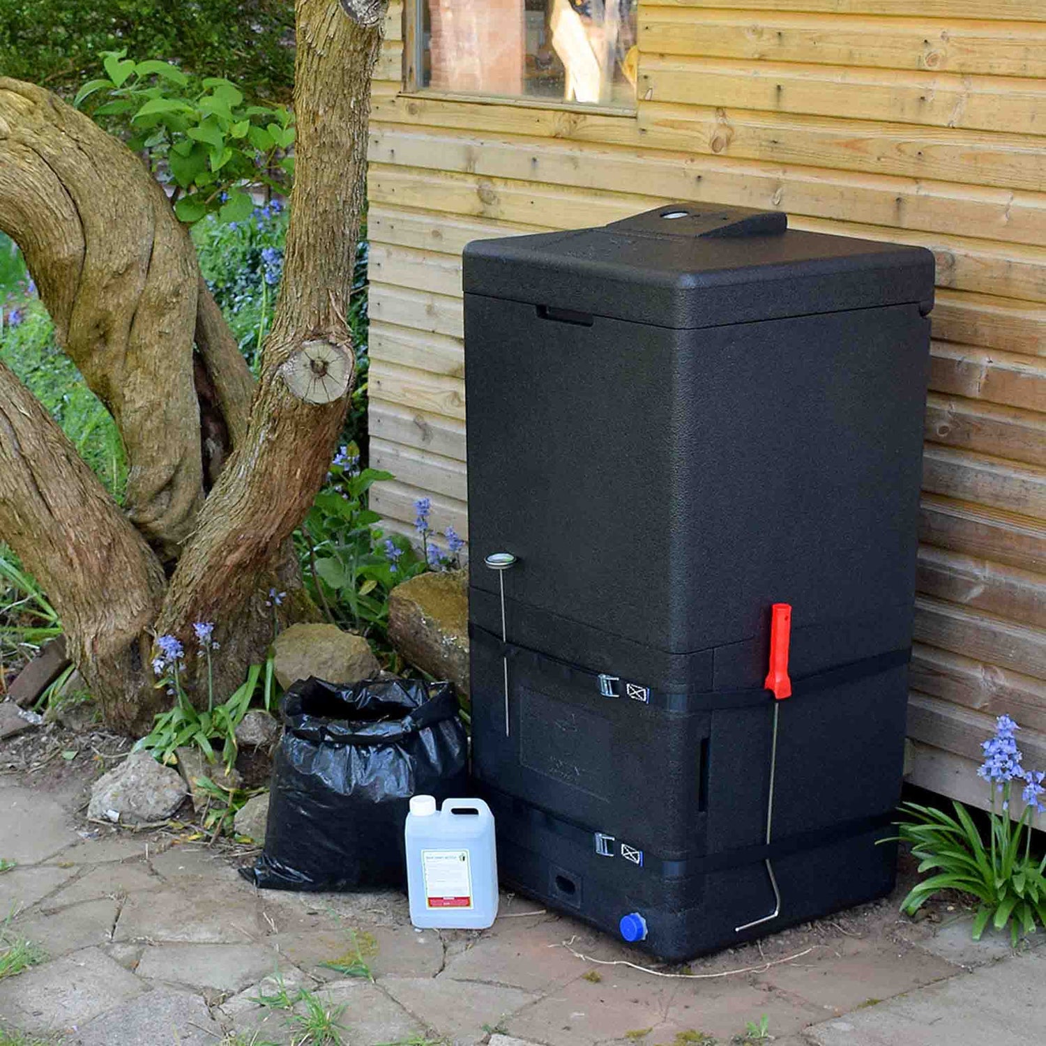 a black HOTBIN 200L composter in a garden by a shed wall